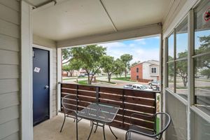 A small balcony with a table and two chairs overlooks a green area with trees. A blue door is visible to the left, while a view of nearby buildings and parked cars can be seen in the background against a partly cloudy sky.