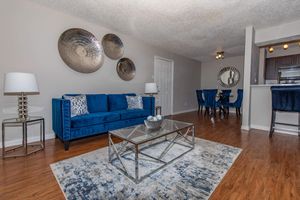 A modern living room featuring a blue velvet sofa with decorative pillows, a glass coffee table on a patterned rug, and circular wall art. The dining area includes a round table with blue chairs, and there’s warm wood flooring throughout the space. Soft lighting adds a cozy ambiance.