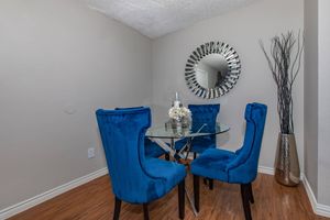 A cozy dining nook featuring a round glass table surrounded by four vibrant blue velvet chairs. A decorative mirror with a sunburst design hangs on the wall, and a tall, slender vase with decorative branches stands nearby. The warm wooden floor adds a touch of elegance to the space.