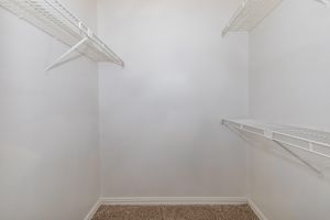 Empty closet with white wire shelves installed on two walls, featuring a light-colored carpeted floor. The walls are plain and white, creating a minimalistic and spacious appearance.