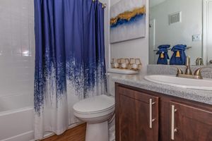 A modern bathroom featuring a white bathtub with a blue gradient shower curtain, a double sink vanity with a granite countertop, a decorative painting on the wall, and neatly arranged gold-colored decor accents. The overall color scheme includes shades of blue, white, and brown.