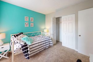 A cozy bedroom featuring a turquoise accent wall, a white bed with black and white striped bedding, a fluffy turquoise throw pillow, and two bedside lamps. There are framed wall art pieces above the bed. A closet with double doors is visible, and the room has a carpeted floor.