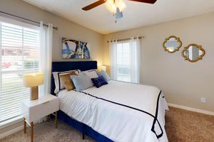 A cozy bedroom featuring a blue upholstered bed with striped bedding, two stylish lamps on bedside tables, and large windows with blinds. Decorative mirrors adorn the wall, and a piece of abstract art adds a modern touch. The room has a warm, inviting atmosphere with soft lighting and carpeted flooring.