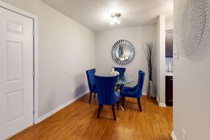 A modern dining area featuring a glass table surrounded by four blue upholstered chairs. The room has light-colored walls, a round mirror with a decorative frame, and a tall decorative plant. The flooring is wooden, and a door is visible on the left side. Soft lighting illuminates the space.