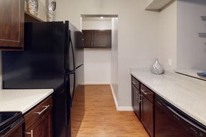 A modern kitchen featuring dark wood cabinets and a black refrigerator. The countertops are light-colored, and the floor has a wood-like finish. A hallway leads to another room in the background, while decorative items are displayed on the shelves. Natural light enhances the space.