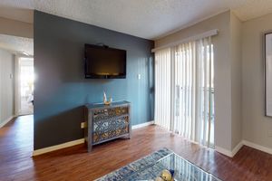 Modern living room with a dark accent wall, featuring a flat-screen TV mounted above a decorative side table. Vertical blinds cover a sliding glass door that leads to a balcony. The floor is hardwood, and a glass coffee table with a decorative item sits in the center of the room. Soft natural light comes through the blinds.