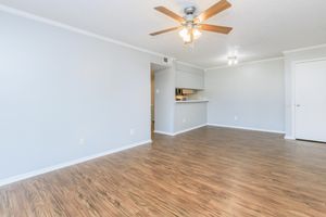 Spacious, empty room with light-colored walls and laminate flooring. A ceiling fan is installed in the center, and a doorway leads to a kitchen area with partial visibility. Natural light brightens the space, creating a calm and inviting atmosphere, ideal for interior design and decoration.