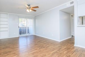 Spacious interior featuring light-colored walls and a ceiling fan. The room has a modern laminate floor, a large window with sliding glass doors leading to a balcony, and built-in shelves on one wall. A doorway to another room is visible on the right side, providing a bright and airy atmosphere.