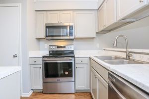 A modern kitchen featuring light gray cabinets, a stainless steel oven and microwave, and a double sink. The countertops are a speckled white with gray marbling. The kitchen layout is compact and includes a doorway leading to another room. The flooring is a warm wood laminate.