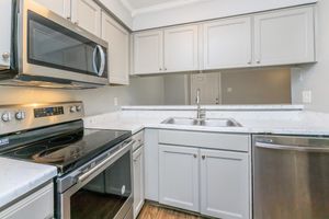 Modern kitchen featuring stainless steel appliances, including a microwave, stove, and dishwasher. The cabinetry is a light gray color, complemented by a white countertop with a marbled design. A double sink is positioned beneath a mirror, enhancing the space's openness. Wood-like flooring completes the contemporary look.