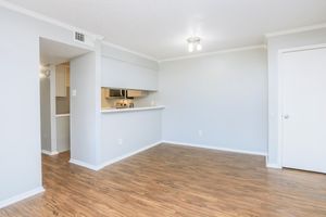 Light-filled interior of a modern apartment featuring grey walls and laminate flooring. The layout includes an open living area transitioning into a kitchen with a bar counter. A closed door leads to another room, and the space is illuminated by a ceiling light fixture.