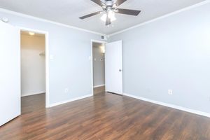 A spacious, empty room with light gray walls and a ceiling fan. Wooden laminate flooring stretches across the floor. Two white doors are visible: one leading to a closet and another to a nearby room, both slightly ajar. The overall atmosphere is bright and inviting, with ample natural light.
