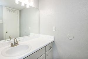 A clean and simple bathroom featuring a white marble countertop with a sink, modern faucet, and vertical mirror. The walls are painted a light gray, and there is a door leading to another room. The space is well-lit with a light fixture above the sink.