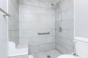 A modern bathroom shower area featuring light gray tiled walls, a handheld showerhead, grab bar for safety, and a built-in shower bench. The floor is tiled with small, round stones, and there is a white toilet adjacent to the shower. The overall design emphasizes a clean and contemporary look.