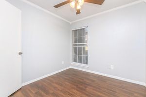 A simple, empty room with light gray walls and hardwood flooring. It features a ceiling fan with light fixtures, a single white door on the left, and a window with white blinds allowing natural light. The space feels bright and open, perfect for various uses.