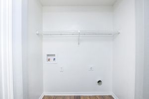 A minimalist empty closet featuring a white wall, a wire shelf mounted on the wall, and a small utility panel with pipes. The floor is wooden, and there is a vent or hole near the baseboard. The overall space appears clean and well-lit.