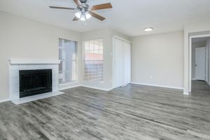 A spacious, well-lit living room featuring a ceiling fan, a modern fireplace with a white surround, large windows with blinds, and a sliding glass door. The flooring is a light-colored laminate, and the walls are painted in a soft neutral tone. There is an adjacent doorway leading to another room.