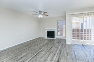 A spacious living room with light-colored walls and wood-look laminate flooring. It features a ceiling fan, a fireplace with a white mantel, and large windows with blinds. A sliding glass door leads to a balcony or patio area. The overall design is modern and inviting, with plenty of natural light.