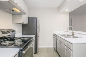 Modern kitchen with a black electric stove, stainless steel refrigerator, and double sink. The countertops are light-colored, and the cabinets are a neutral shade, creating a clean and spacious look. The floor is tiled in a light gray color, enhancing the kitchen's overall brightness.
