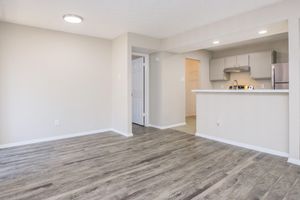 A spacious, well-lit living area with light-colored walls and wooden-style flooring. A small white bar separates the living space from a kitchen in the background. There's a door leading to another room, and the overall atmosphere is clean and modern.