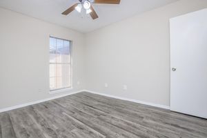 A light-colored room featuring a ceiling fan, large window with blinds, and hardwood-style flooring. The walls are painted in a neutral tone, and there is a closed white door on the right. The room is empty, providing a spacious feel.