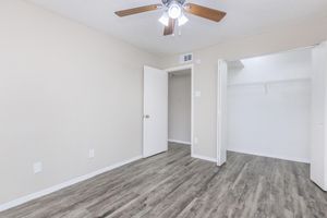 A light-colored room with a ceiling fan, featuring beige walls and a wooden floor. There are two white doors, one leading to an empty closet with a rod for hangers. The room has ample natural light, creating a bright and airy atmosphere.