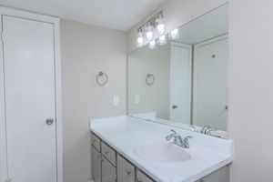 A bathroom interior featuring a white countertop with a sink, a large mirror above, and four light fixtures. The walls are painted in a neutral tone, and there is a towel ring on the wall. A closed door is visible on the left side of the image.
