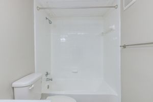A clean, minimalistic bathroom featuring a white bathtub with a showerhead, a white toilet, and a simple wall-mounted towel rack. The walls and tiles are light-colored, contributing to a bright and airy feel. The space is uncluttered and designed for practicality.