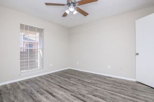 Empty room with light-colored walls, a ceiling fan with wooden blades, and a window with blinds allowing natural light. The floor is covered in laminate wood, and there is a white door on the right side. The space appears clean and minimalistic, suitable for various uses.