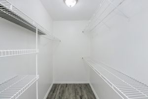 A spacious, empty closet featuring white wire shelving on both sides and a light fixture on the ceiling. The walls are painted white, and the floor has a wood-like design, creating a clean and bright storage area.