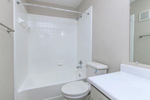 A clean, empty bathroom featuring a white bathtub with a shower curtain, a toilet, and a sink with a countertop. The walls are light gray, and there is a mirror above the sink. The space appears modern and well-maintained.