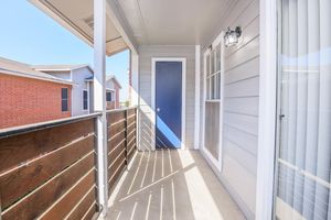 A view of a balcony with a blue door and wooden railing. Sunlight casts shadows on the concrete floor, highlighting the wooden planks of the railing. In the background, there are several residential buildings, including brick and light-colored houses, under a clear blue sky.