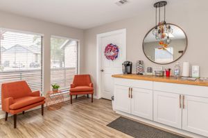 Cozy waiting area featuring two orange chairs, a circular mirror, and a wooden counter with coffee supplies. A red and blue wreath adorns the wall, while natural light streams in through the windows with blinds. A small plant and various brochures are visible on the counter.