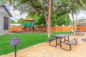 A grassy outdoor area featuring a playground with slides and climbing structures, surrounded by a wooden fence. In the foreground, there are two black picnic tables and a charcoal grill, with a large tree providing shade. The sky is partly cloudy, creating a bright and inviting atmosphere.