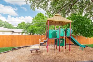Colorful children's playground equipped with slides, climbing structures, and play panels. It sits on a bed of wood chips, surrounded by a wooden fence and green trees, under a partly cloudy sky. The playground invites outdoor play and exploration.