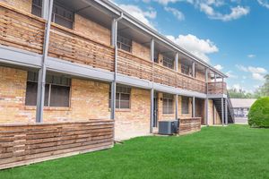 A two-story brick building with wooden balcony railings and grassy landscaping. The structure features multiple windows and a staircase on one side, against a backdrop of a blue sky with scattered clouds.