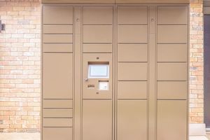 A tan delivery locker unit against a brick wall, featuring a digital screen and buttons for package access. The locker is designed for secure package retrieval, with multiple compartments for different sizes of deliveries.