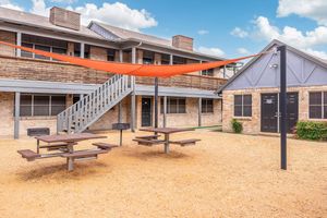 An outdoor area featuring several picnic tables shaded by an orange canopy. In the background, a two-story building with a wooden facade and multiple doors is visible. The ground is covered in gravel, and a staircase leads to the upper level of the building. Blue skies with fluffy clouds can be seen above.