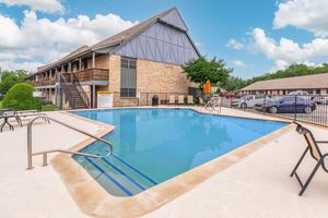 A clear swimming pool surrounded by lounge chairs and an orange umbrella, located next to a multi-story brick building. Lush greenery is visible, and parked cars are in the background under a bright blue sky with fluffy white clouds.