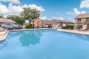 A serene swimming pool surrounded by a patio area with chairs and umbrellas. Nearby, there are trees and several buildings in the background. The sky is partly cloudy, creating a bright, inviting atmosphere for relaxation and leisure.