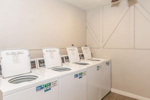 A clean laundry room featuring four white top-loading washing machines, each equipped with control panels. The room has light-colored walls and a ceiling fan, creating a bright and spacious environment.