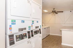 Self-service laundry area featuring several stacked washing machines and dryers with digital control panels. A ceiling fan provides airflow in the room, which has a clean and modern design with light-colored walls and wood-style flooring.