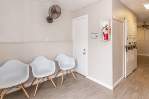 A minimalist waiting area featuring three white chairs with wooden legs, a wall-mounted fire extinguisher, a "No Smoking" sign, and a laundry area with multiple machines. The walls are painted a light color, with a ceiling fan visible in the background.