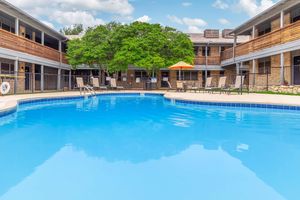 A bright, inviting swimming pool surrounded by lounge chairs and an orange umbrella, set within a courtyard of a two-story building. Lush greenery and a clear blue sky create a relaxed atmosphere, perfect for relaxation and leisure.