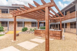 A rustic wooden pergola with slatted roofing stands in a courtyard, featuring a wooden service counter and stone paths. Surrounding the area are low-maintenance plants and gravel, complemented by an inviting atmosphere of a contemporary building with brick walls and large windows.