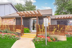 A welcoming entrance to an apartment complex featuring a wooden pergola, colorful flowers, and a sign for future resident parking. The building has a combination of brick and wooden elements, with large windows and a well-maintained lawn. Lush greenery and landscaping enhance the inviting atmosphere.