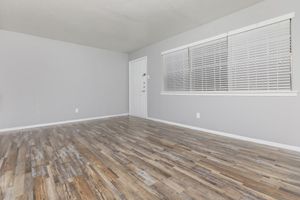 A spacious room with light gray walls and a wooden floor. It features a white door leading outside and a window with horizontal blinds. The space is empty, creating a minimalistic and airy atmosphere, ideal for various interior design options.