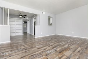 Spacious interior of a light gray living room with wooden flooring, featuring an open layout. A ceiling fan is visible, along with a shelf in the background. Natural light enters through a window, enhancing the airy atmosphere. The room is unadorned and ready for decoration or furniture placement.