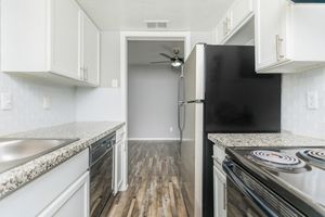 Modern kitchen featuring white cabinets, a granite countertop, and stainless steel appliances. A black refrigerator and an oven with a stove are visible. The background shows a doorway leading to a room with a ceiling fan and light gray walls, indicating an open floor plan.