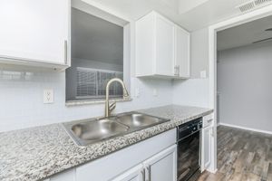 A modern kitchen with a granite countertop featuring a double sink, a stainless steel faucet, and white cabinetry. The backsplash is made of light blue tiles. A window above the sink lets in natural light, and there is a glimpse of a room with wooden flooring in the background.
